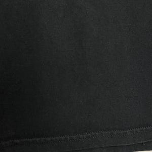 May include: Close-up of a black fabric, likely a t-shirt or similar garment. The image shows the texture of the material and the stitched hem at the bottom. The fabric appears to be a solid, dark color.