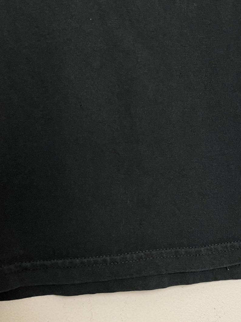May include: Close-up of a black fabric, likely a t-shirt or similar garment. The image shows the texture of the material and the stitched hem at the bottom. The fabric appears to be a solid, dark color.