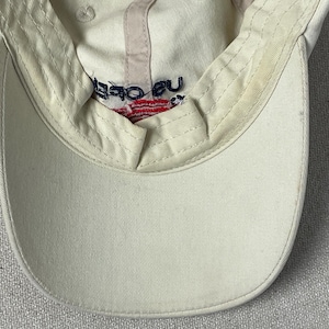 May include: An off-white baseball cap featuring an interior sweatband with visible signs of wear. The inner crown displays embroidered text reading US OPEN in dark blue, positioned above a small, red and white striped graphic element.
