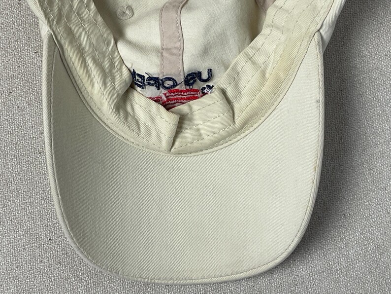 May include: An off-white baseball cap featuring an interior sweatband with visible signs of wear. The inner crown displays embroidered text reading US OPEN in dark blue, positioned above a small, red and white striped graphic element.