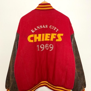 May include: A red vintage letterman jacket with brown leather sleeves. The back displays the words "Kansas City Chiefs" in yellow and white, with "1969" below. The collar and cuffs have yellow and red stripes.