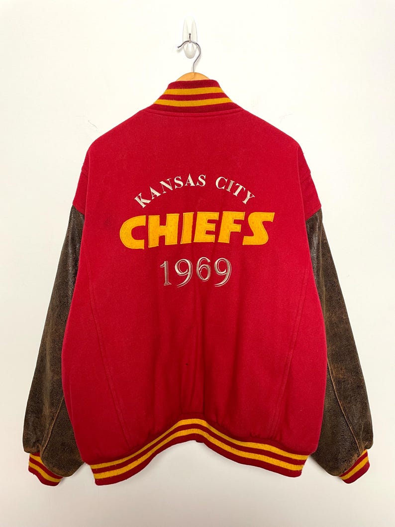 May include: A red vintage letterman jacket with brown leather sleeves. The back displays the words "Kansas City Chiefs" in yellow and white, with "1969" below. The collar and cuffs have yellow and red stripes.