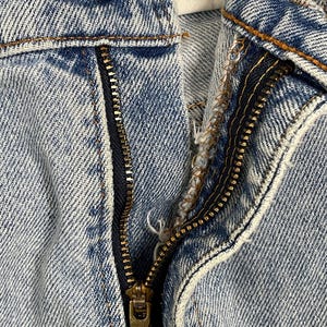 May include: Close-up of light blue denim jeans with a partially unzipped brass zipper. A brass button is visible at the top, along with orange stitching. The fabric shows signs of wear and tear.
