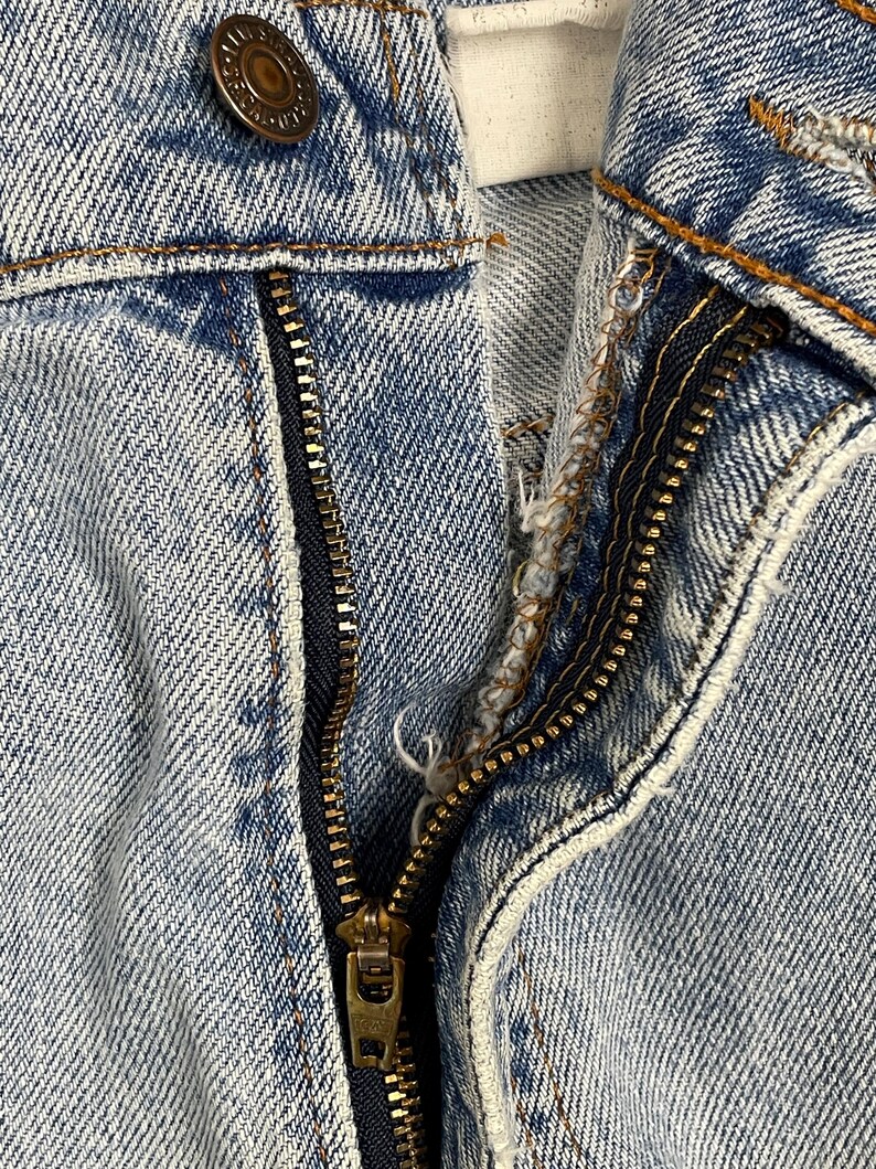 May include: Close-up of light blue denim jeans with a partially unzipped brass zipper. A brass button is visible at the top, along with orange stitching. The fabric shows signs of wear and tear.