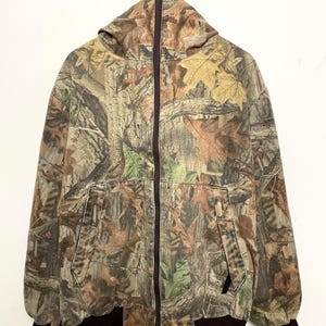 Puede incluir: Chaqueta con capucha de camuflaje con cremallera completa. La chaqueta presenta un estampado de hojas marrones y verdes, con un dobladillo y puños acanalados de color marrón oscuro. Tiene dos bolsillos delanteros y una capucha.
