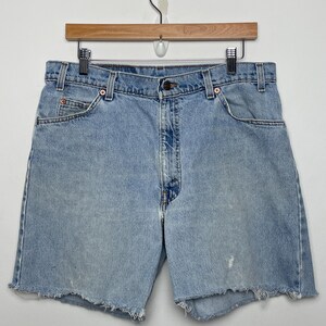 May include: Light blue denim shorts with a frayed hem, hanging on a wooden hanger. The shorts have a classic five-pocket design and a button closure. The denim has a faded appearance.