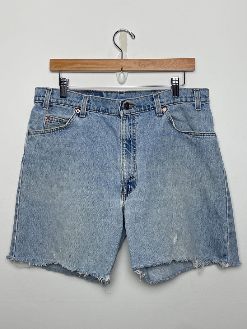 May include: Light blue denim shorts with a frayed hem, hanging on a wooden hanger. The shorts have a classic five-pocket design and a button closure. The denim has a faded appearance.