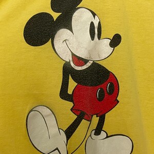 K&ouml;nnte beinhalten: Gelbes T-Shirt mit einer Mickey Mouse-Illustration. Die Zeichentrickfigur ist in Schwarz, Wei&szlig; und Rot gehalten und l&auml;chelt. Der Text "Walt Disney Productions" ist unten im Bild aufgedruckt.
