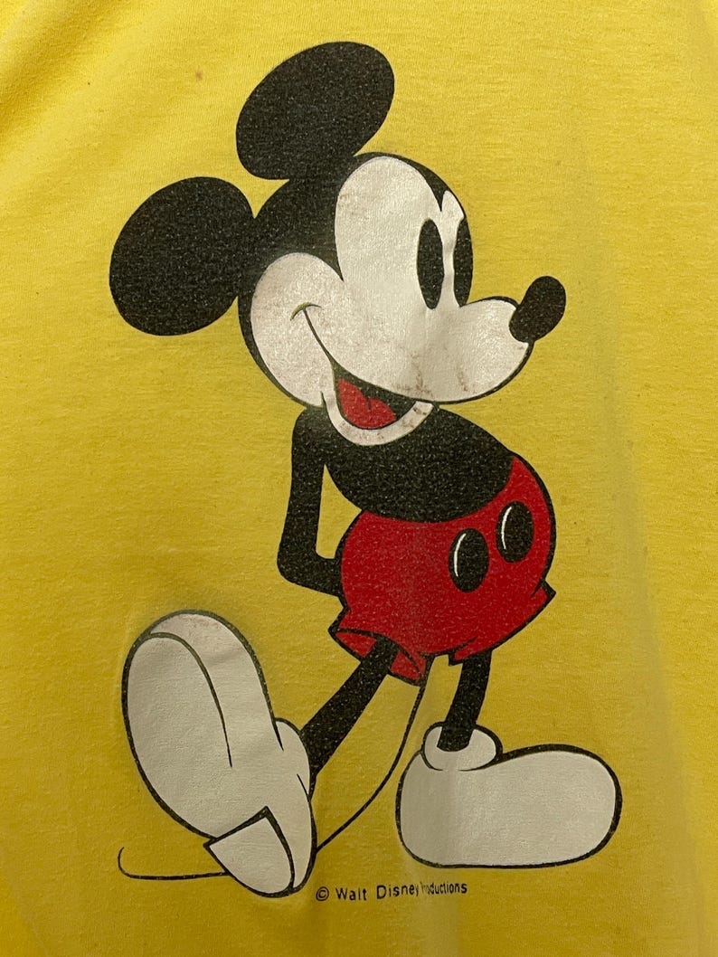K&ouml;nnte beinhalten: Gelbes T-Shirt mit einer Mickey Mouse-Illustration. Die Zeichentrickfigur ist in Schwarz, Wei&szlig; und Rot gehalten und l&auml;chelt. Der Text "Walt Disney Productions" ist unten im Bild aufgedruckt.