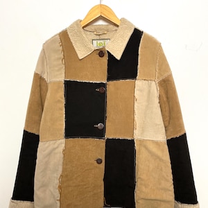 May include: A patchwork jacket in shades of tan, beige, and black, with a cream-colored shearling collar and cuffs. The jacket has a button-up front and is hanging on a wooden hanger.