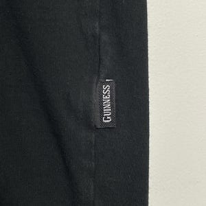 May include: Close-up of black fabric with a small, rectangular label. The label reads "GUINNESS" in white lettering, suggesting a branded clothing item. The fabric is likely part of a t-shirt or similar garment.