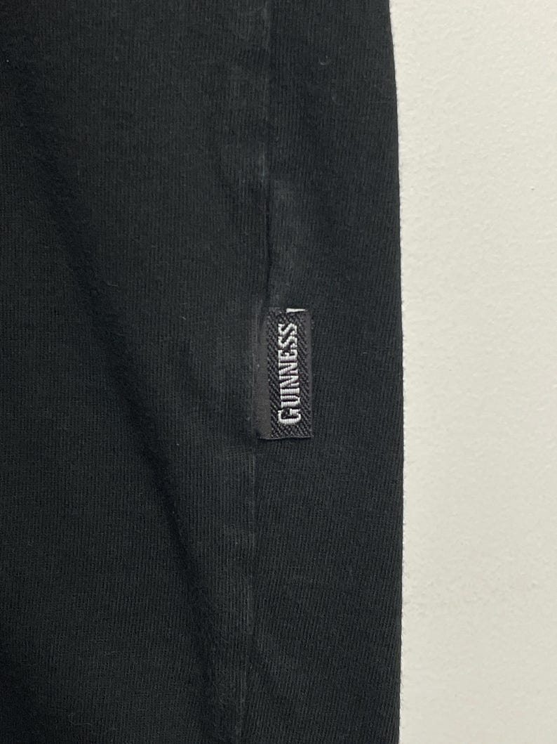 May include: Close-up of black fabric with a small, rectangular label. The label reads "GUINNESS" in white lettering, suggesting a branded clothing item. The fabric is likely part of a t-shirt or similar garment.