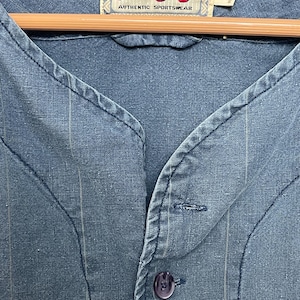 May include: A blue button-down shirt with thin vertical pinstripes and a V-neckline. The interior neck label reads Mirage First String Authentic Sportswear. The shirt features a single visible dark blue button and textured fabric construction.