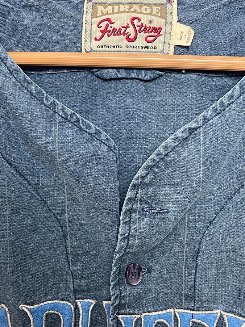 May include: A blue button-down shirt with thin vertical pinstripes and a V-neckline. The interior neck label reads Mirage First String Authentic Sportswear. The shirt features a single visible dark blue button and textured fabric construction.