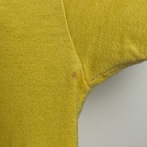 K&ouml;nnte beinhalten: Nahaufnahme eines leuchtend gelben T-Shirts, das die Textur des Stoffes zeigt. Das Bild konzentriert sich auf den Schulter- und &Auml;rmelbereich, mit einem kleinen Fleck in der N&auml;he der Achselh&ouml;hle. Der Hintergrund ist schlicht wei&szlig;.