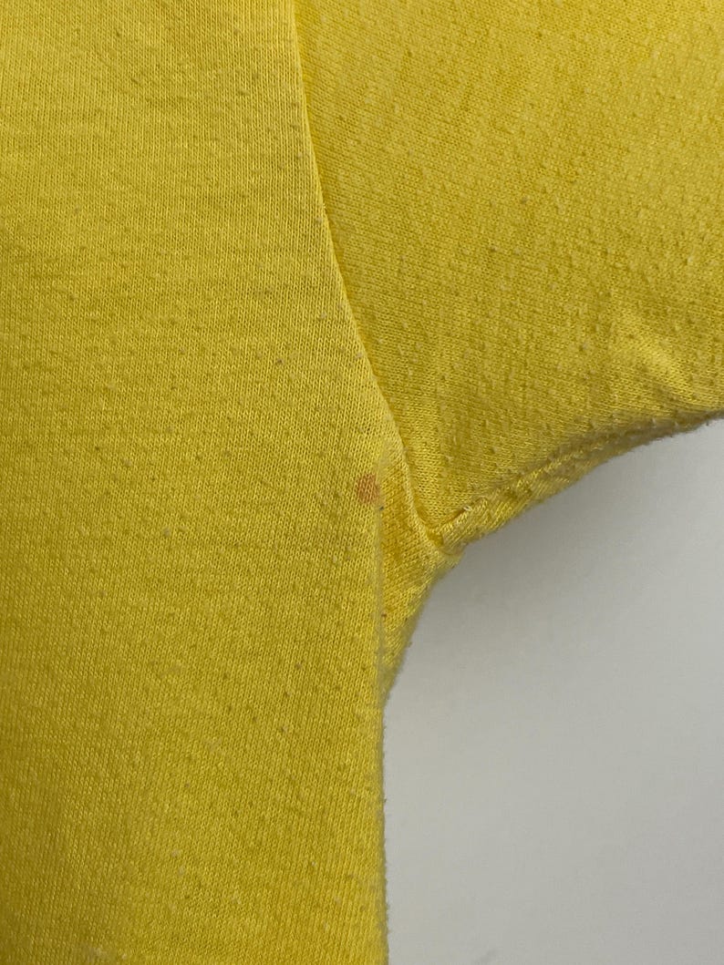 K&ouml;nnte beinhalten: Nahaufnahme eines leuchtend gelben T-Shirts, das die Textur des Stoffes zeigt. Das Bild konzentriert sich auf den Schulter- und &Auml;rmelbereich, mit einem kleinen Fleck in der N&auml;he der Achselh&ouml;hle. Der Hintergrund ist schlicht wei&szlig;.