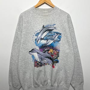 Puede incluir: Sudadera gris con un gráfico de tres delfines saltando sobre corales coloridos. La palabra "SEAWORLD" está impresa en un círculo alrededor de los delfines. La sudadera está colgada en una percha de madera.