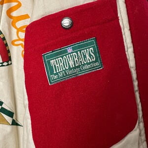 May include: Detail of a red pocket on a vintage jacket. The pocket displays a green and white label reading "THROWBACKS The NFL Vintage Collection." A silver button with a football design is above the pocket. The jacket's interior is cream-coloured.