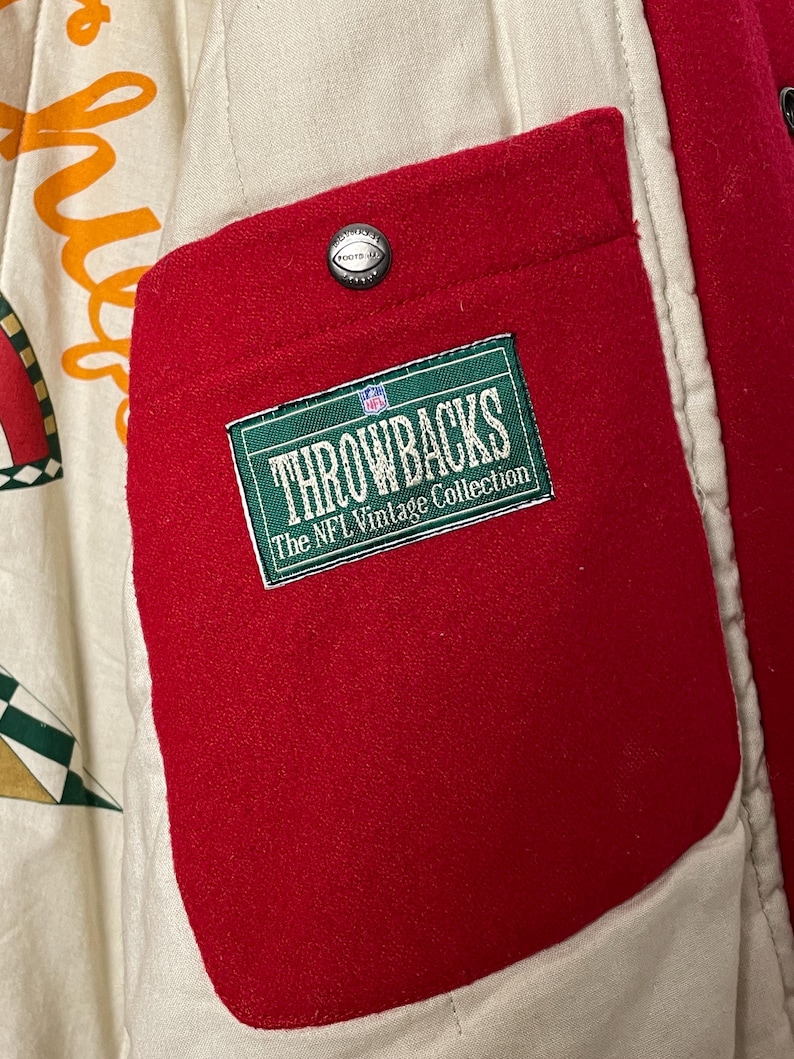 May include: Detail of a red pocket on a vintage jacket. The pocket displays a green and white label reading "THROWBACKS The NFL Vintage Collection." A silver button with a football design is above the pocket. The jacket's interior is cream-coloured.