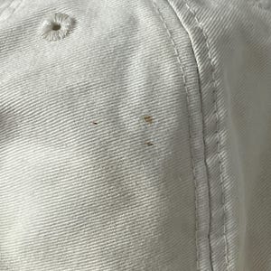 May include: A white baseball cap made of cotton twill fabric featuring stitched eyelets for ventilation and reinforced seams. The surface of the cap displays three small, distinct brown spots located on the side panel near the vertical seam.