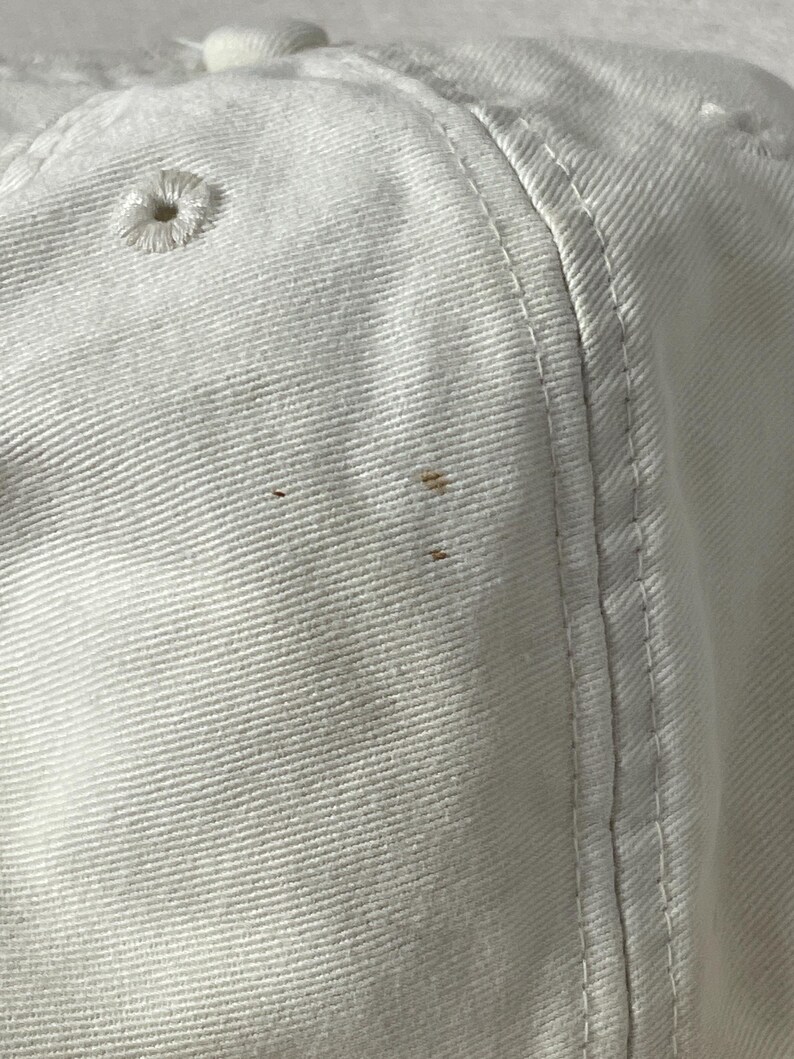 May include: A white baseball cap made of cotton twill fabric featuring stitched eyelets for ventilation and reinforced seams. The surface of the cap displays three small, distinct brown spots located on the side panel near the vertical seam.