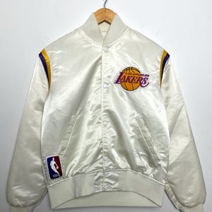 Puede incluir: Chaqueta de satén color crema con el logotipo de Los Angeles Lakers en morado y dorado. Cuenta con cuello, puños y dobladillo de canalé blanco. Incluye rayas moradas y doradas en los hombros y un parche con el logo de la NBA.