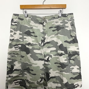 May include: Camouflage cargo shorts in shades of green, gray, and white, hanging on a wooden hanger. The shorts feature multiple pockets and a button closure. The fabric has a classic military-inspired pattern.