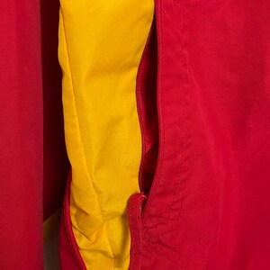Puede incluir: Primer plano de una chaqueta roja y amarilla. La chaqueta tiene un exterior rojo y un forro interior amarillo. Una cremallera roja es visible en el lado derecho. La chaqueta parece estar hecha de un material liso, posiblemente nailon.