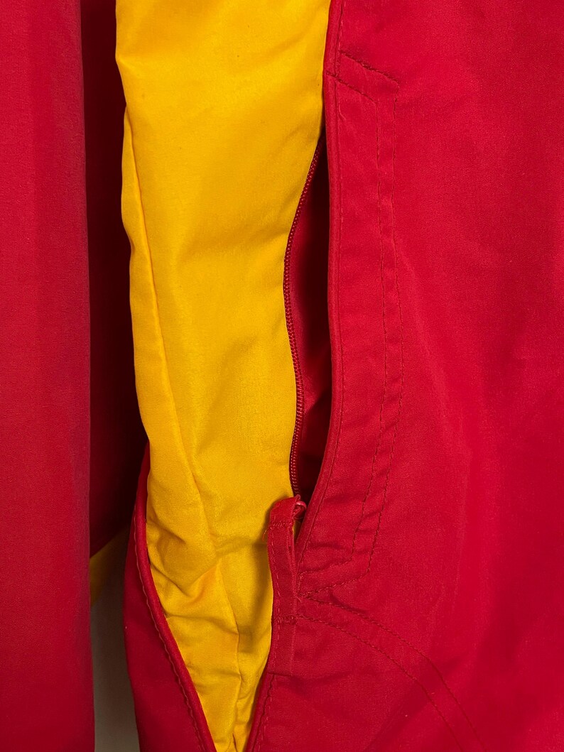 Puede incluir: Primer plano de una chaqueta roja y amarilla. La chaqueta tiene un exterior rojo y un forro interior amarillo. Una cremallera roja es visible en el lado derecho. La chaqueta parece estar hecha de un material liso, posiblemente nailon.