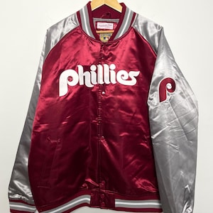 May include: A maroon and silver satin baseball jacket with the word "Phillies" in white script. The sleeves are silver, and the jacket has a striped collar, cuffs, and hem. A "P" logo is on the left sleeve. The jacket is hanging on a hook.