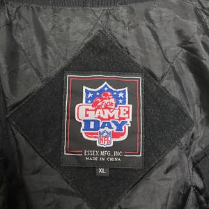 Pode incluir: Casaco preto acolchoado com gola listrada verde e preta. O casaco apresenta um patch da NFL "Game Day" com um gr&aacute;fico de jogador de futebol. A etiqueta diz "Essex Mfg, Inc. Made in China" e &eacute; tamanho XL.