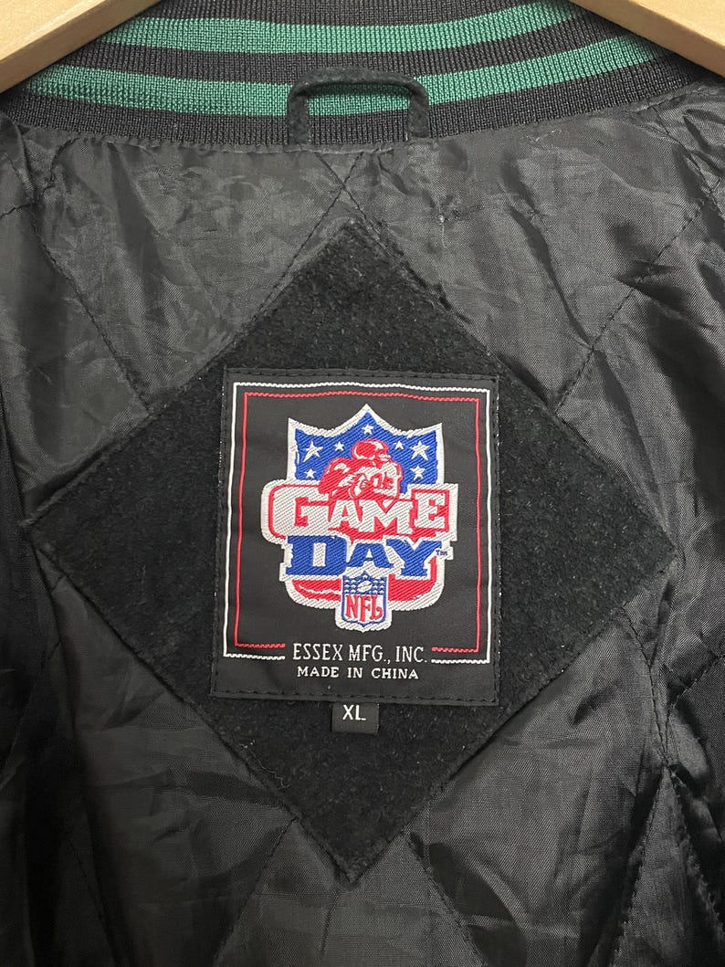 Pode incluir: Casaco preto acolchoado com gola listrada verde e preta. O casaco apresenta um patch da NFL "Game Day" com um gr&aacute;fico de jogador de futebol. A etiqueta diz "Essex Mfg, Inc. Made in China" e &eacute; tamanho XL.