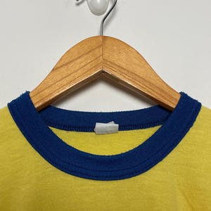 K&ouml;nnte beinhalten: Nahaufnahme eines gelben T-Shirts mit blauem Kragen. Das Shirt h&auml;ngt an einem Holzkleiderb&uuml;gel vor einer wei&szlig;en Wand. Der Kragen ist dunkelblau und bildet ein kontrastreiches Design.
