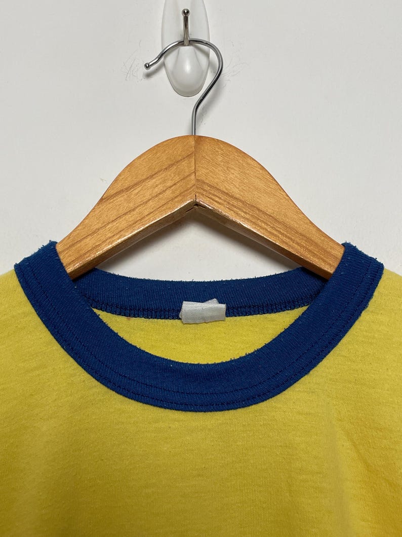 K&ouml;nnte beinhalten: Nahaufnahme eines gelben T-Shirts mit blauem Kragen. Das Shirt h&auml;ngt an einem Holzkleiderb&uuml;gel vor einer wei&szlig;en Wand. Der Kragen ist dunkelblau und bildet ein kontrastreiches Design.