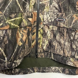 Pode incluir: Casaco de ca&ccedil;a camuflado com z&iacute;per preto de comprimento total e dois bolsos frontais. O casaco apresenta um padr&atilde;o de cores castanho, verde e bege, com um pequeno patch que diz "SURVIVMAX".