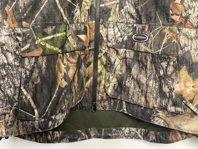 Pode incluir: Casaco de ca&ccedil;a camuflado com z&iacute;per preto de comprimento total e dois bolsos frontais. O casaco apresenta um padr&atilde;o de cores castanho, verde e bege, com um pequeno patch que diz "SURVIVMAX".