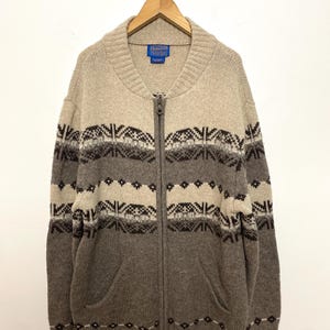 Puede incluir: Cárdigan con cremallera en tonos beige, marrón y gris, con un patrón geométrico en el pecho y las mangas. El suéter tiene un cuello alto y dos bolsillos delanteros. La etiqueta de la marca es visible en el cuello.