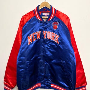 Peut inclure: Une veste de baseball en satin bleu et rouge avec les mots "New York" et un logo de basket-ball sur le devant. La veste a des rayures blanches et rouges sur les manches et le bas.