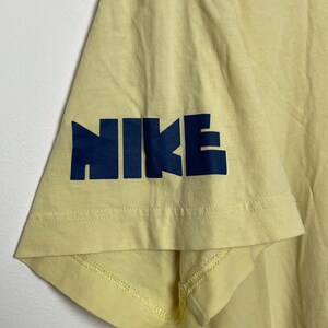 May include: Close-up of a pale yellow t-shirt sleeve. The word "NIKE" is printed in navy blue block letters on the sleeve. The shirt appears to be made of a soft, breathable fabric.