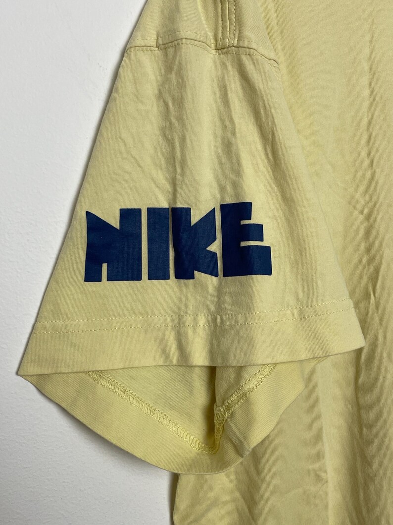 May include: Close-up of a pale yellow t-shirt sleeve. The word "NIKE" is printed in navy blue block letters on the sleeve. The shirt appears to be made of a soft, breathable fabric.
