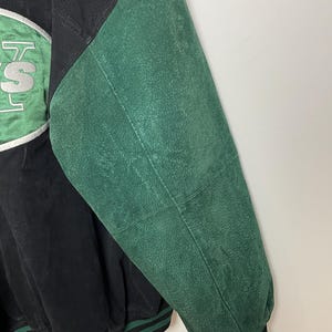 Pode incluir: Casaco vintage preto e verde com um patch com o log&oacute;tipo da equipa. O casaco apresenta um corpo preto com uma manga verde e uma guarni&ccedil;&atilde;o listrada verde e preta. O log&oacute;tipo da equipa &eacute; em branco e verde.