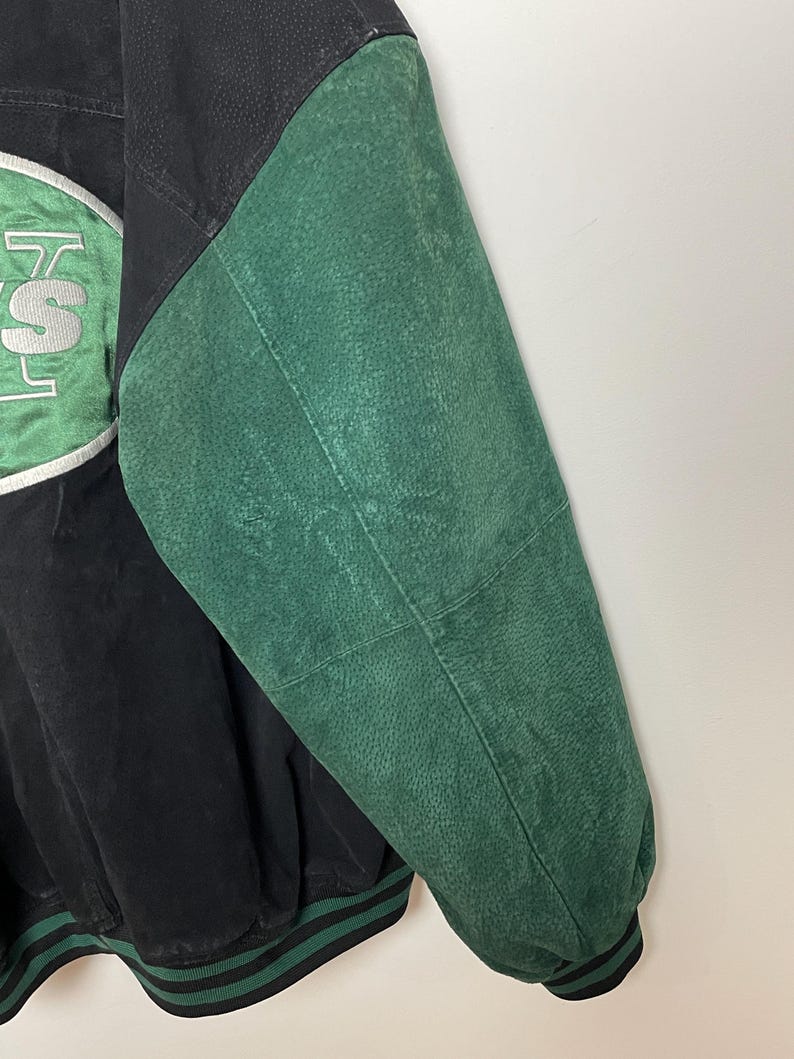 Pode incluir: Casaco vintage preto e verde com um patch com o log&oacute;tipo da equipa. O casaco apresenta um corpo preto com uma manga verde e uma guarni&ccedil;&atilde;o listrada verde e preta. O log&oacute;tipo da equipa &eacute; em branco e verde.