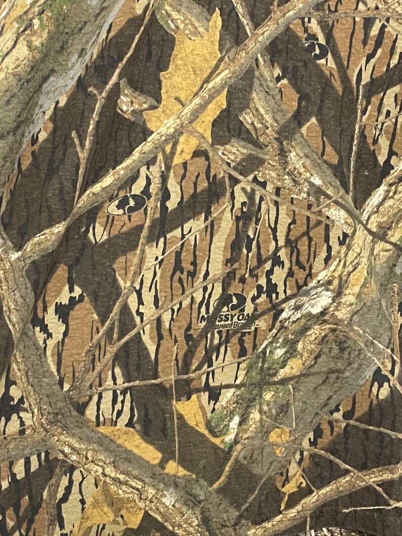 May include: Close-up of a camouflage pattern in shades of brown, tan, and green. The design includes tree branches and leaf shapes, with the "Mossy Oak" logo visible.