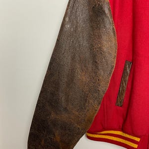 May include: Red varsity jacket with brown leather sleeves. The jacket features a yellow and red striped cuff and a brown leather pocket detail. The leather sleeves have a distressed appearance.
