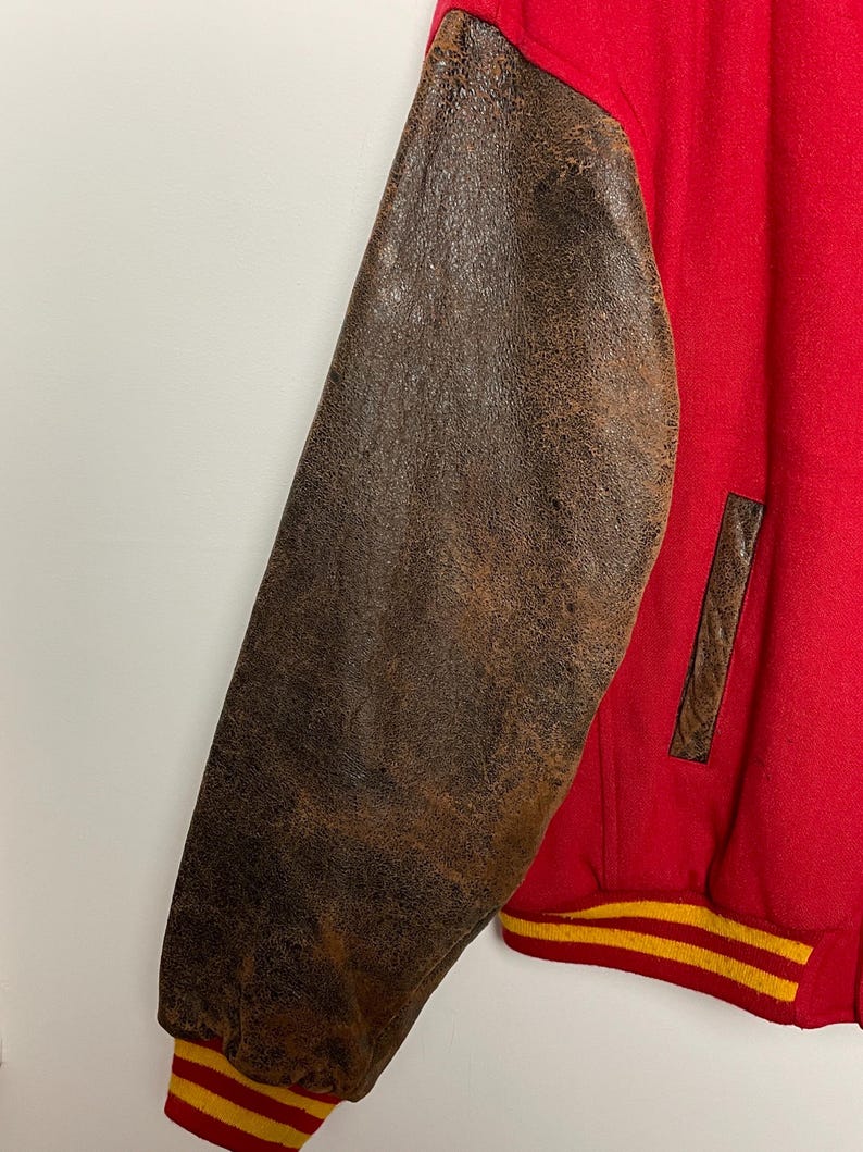 May include: Red varsity jacket with brown leather sleeves. The jacket features a yellow and red striped cuff and a brown leather pocket detail. The leather sleeves have a distressed appearance.