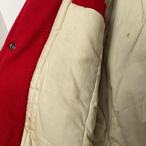 May include: Close-up of a red wool jacket with a cream quilted lining. The jacket features a button closure and a red accent on the inside. The lining shows some stains.