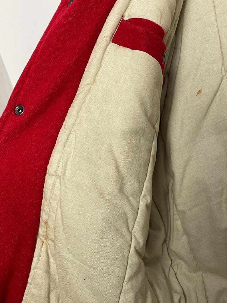 May include: Close-up of a red wool jacket with a cream quilted lining. The jacket features a button closure and a red accent on the inside. The lining shows some stains.