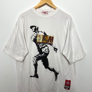Peut inclure: T-shirt blanc avec une silhouette noire d'un joueur de baseball tenant une batte. Le joueur porte une casquette et le t-shirt a un graphique d'une carte de baseball avec le texte "M" dessus. Le t-shirt a une étiquette rouge avec le texte "XL" dessus.