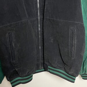 Pode incluir: Casaco preto com detalhes verdes. O casaco tem fecho de correr, dois bolsos frontais e punhos listrados em verde e preto. As letras "NYJ" s&atilde;o bordadas na parte superior direita do casaco.