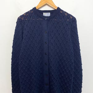 May include: A navy blue long-sleeve cardigan featuring a textured diamond knit pattern with raised bobbles. The garment has a round neckline, a front button closure with matching dark buttons, and a scalloped hemline. A label is visible at the neck.
